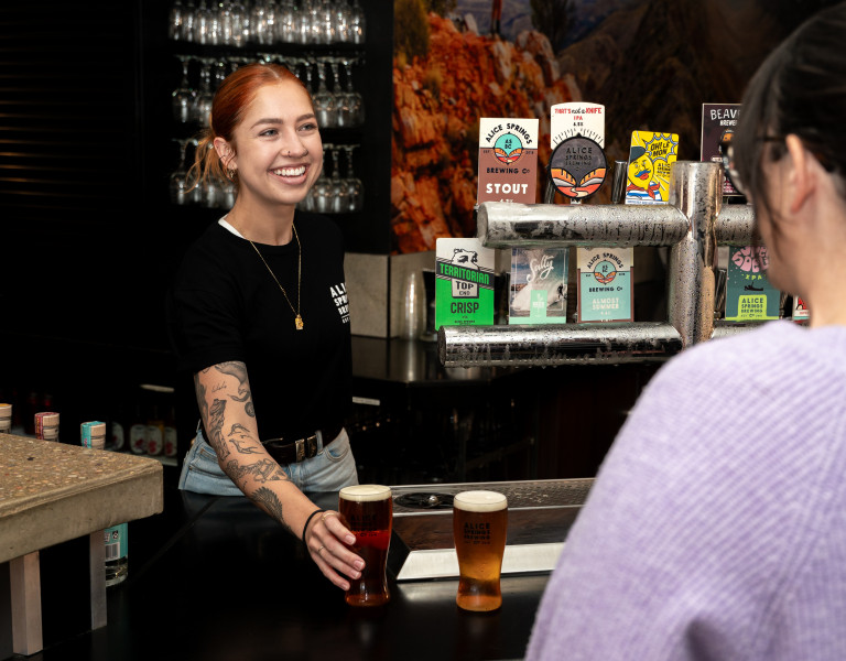 A bartender smiling as she serves drinks across the bar, with beer taps, menus and bar décor behind her.