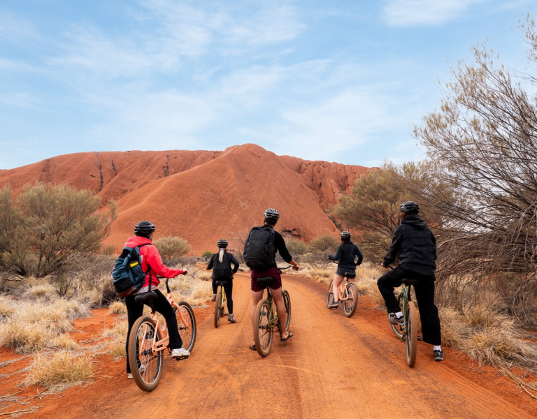 Four cyclists riding along a red dirt track surrounded by scrub and low trees, with distant rocky hills under a partly cloudy sky.