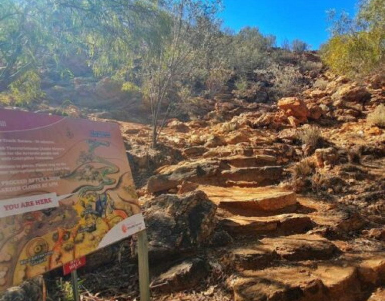 A rocky walking trail leading uphill through dry bushland, with a colourful information sign in the foreground.