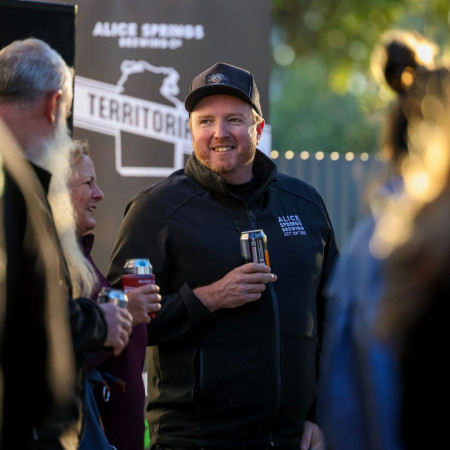 A man smiling while holding a beer can at an outdoor event, with people chatting around him and an Alice Springs Brewing Co sign in the background.