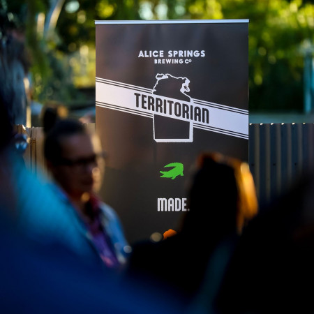A blurred crowd in the foreground with a focus on an Alice Springs Brewing Co branded backdrop at an event.