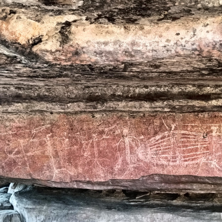 Ancient Aboriginal rock art painted on a rock wall, featuring faded figures and patterns in earthy red and white tones.
