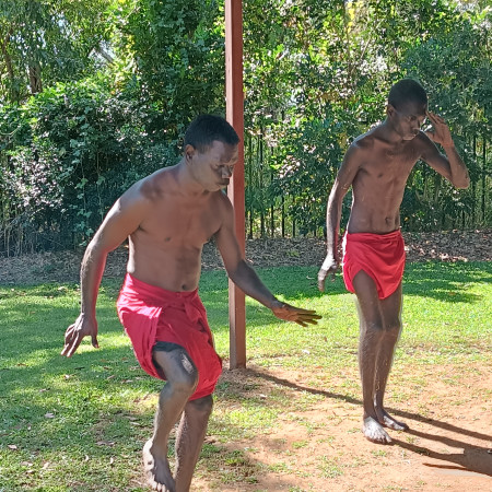 Two Aboriginal men performing a traditional dance outdoors, both barefoot and wearing red shorts.