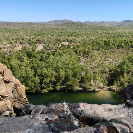 A wide landscape view of rocky terrain and dense green bushland surrounding a calm waterhole beneath a cliff.