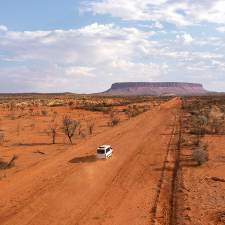 A white 4WD driving along a red dirt road in the outback, with Mt Conner visible in the distance under a partly cloudy sky.