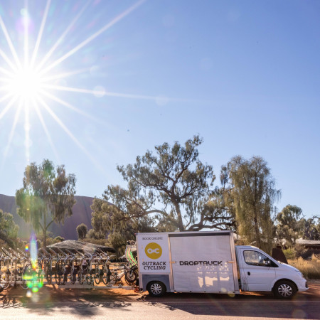 Cyclists gathering beside a support vehicle in the outback, with tall trees and hills under a bright sunburst in a clear blue sky.