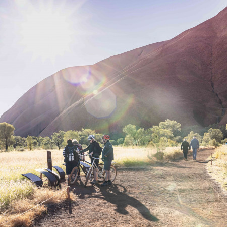 A group of cyclists preparing their bikes at the base of Uluru, with soft morning light and long shadows cast across the desert landscape.