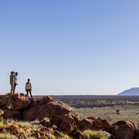 Three people standing on a rocky outcrop overlooking a vast desert landscape at sunset, with long shadows cast across the ground.