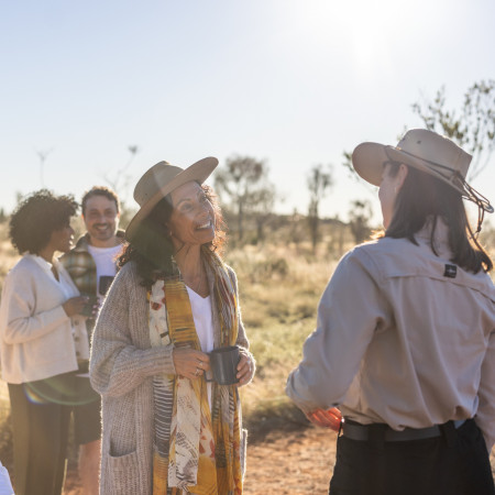 A small tour group chatting outdoors in warm light, with a woman in a hat holding a cup and others gathered casually around her.
