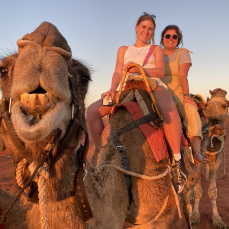 Two people sit smiling on a camel during a sunset camel ride in the desert. A close-up of the camel's face dominates the foreground, giving the image a fun, friendly feel.