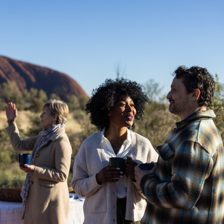 Three people talking and smiling during sunrise at Uluru, dressed warmly and standing in soft golden light.
