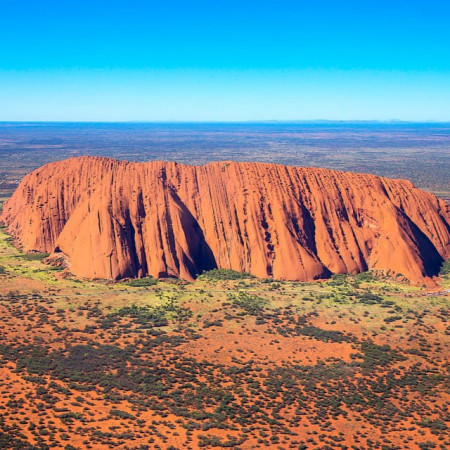 Aerial view of Uluru rising from red desert landscape under clear blue sky in Central Australia.