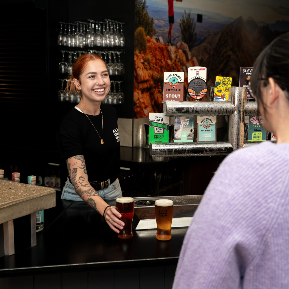 A bartender smiling as she serves drinks across the bar, with beer taps, menus and bar décor behind her.